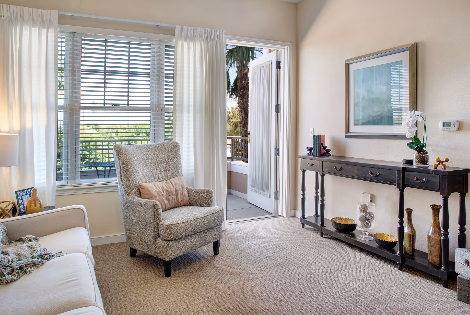 Bright living room with an armchair by a large window, a sofa, and a console table against the wall with a door opening to a small balcony.