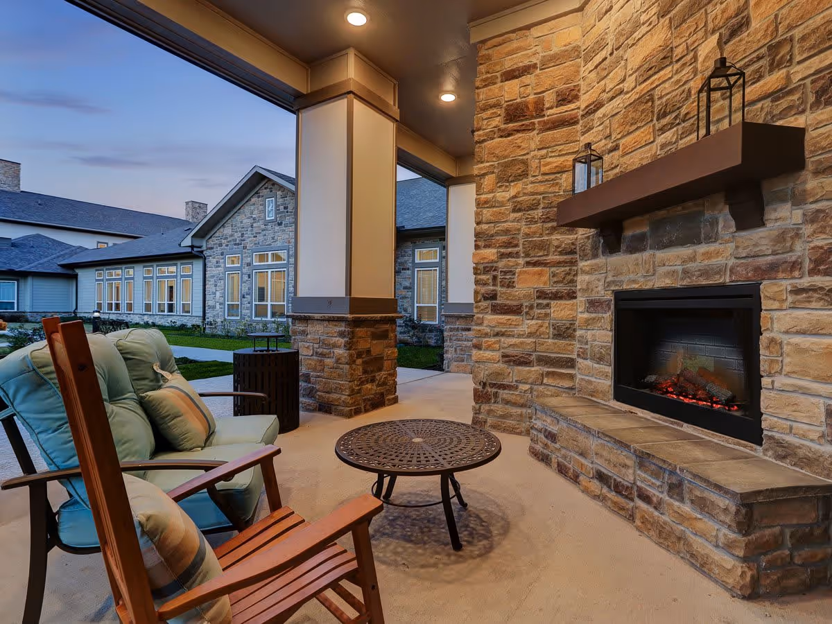 Covered outdoor patio area with cushioned wooden chairs and a round metal table in front of a stone fireplace with a wooden mantel holding two lanterns. The patio overlooks a well-maintained lawn and a stone building with multiple windows during dusk.