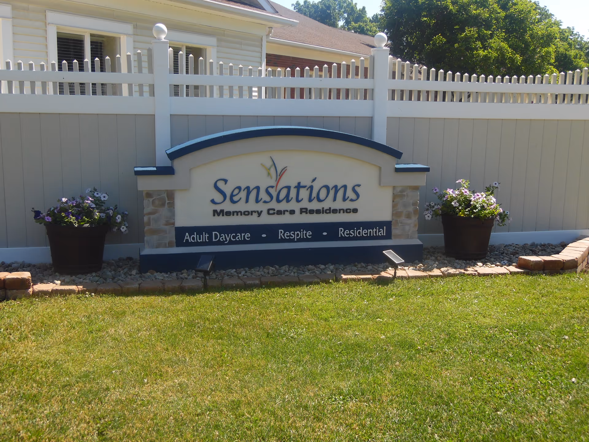 Outdoor sign reading "Sensations Memory Care Residence" in front of a white fence with potted flowers and a grassy lawn.