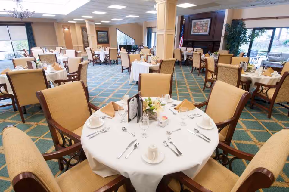 A spacious dining room with multiple round tables covered with white tablecloths, each set with plates, cups, silverware, napkins, and small floral centerpieces. The room has beige upholstered chairs, large windows letting in natural light, and a carpeted floor with a green and gold pattern.