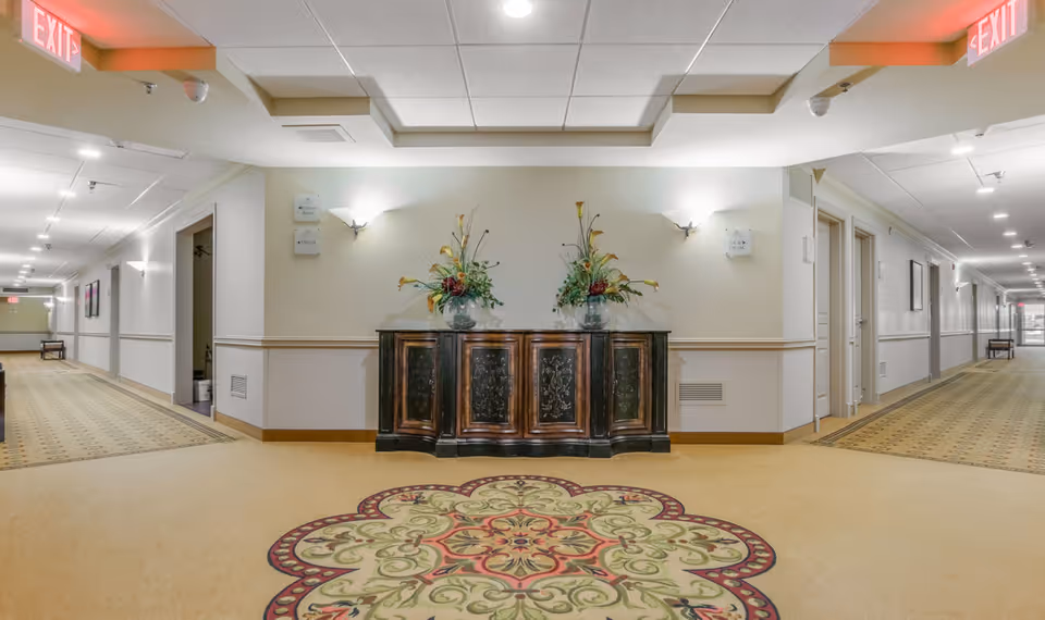 Carpeted senior-living hallway with a decorative console table topped with floral arrangements at a central vestibule and corridors extending left and right.