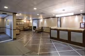 Interior view of a reception area in a senior living facility with a front desk on the right, several chairs along the walls, and a hallway leading to other rooms. The floor has a tiled pattern and the ceiling has recessed lighting.