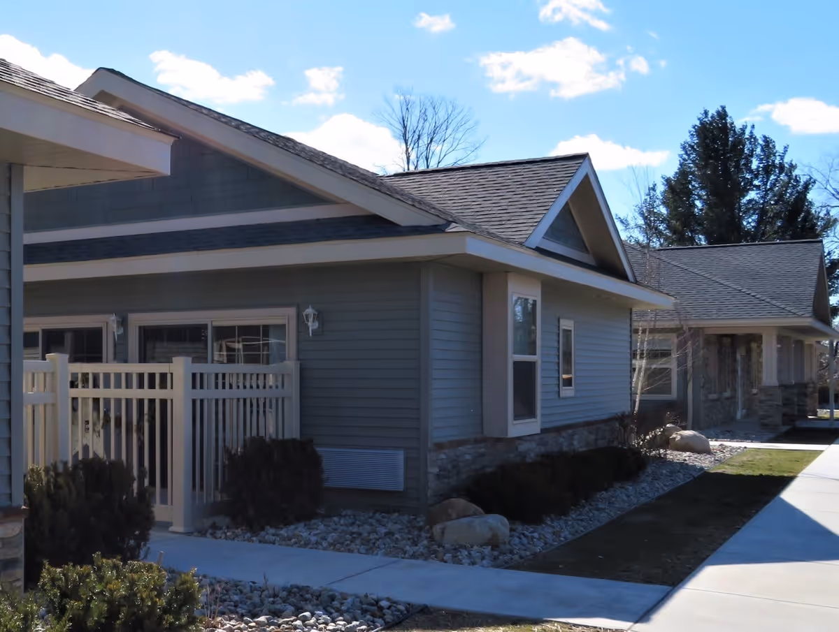 Exterior view of a single-story residential building with gray siding, white trim, and a shingled roof under a blue sky with some clouds. There is a small fenced patio area and landscaped rocks and bushes along the building. A concrete sidewalk runs alongside the building.