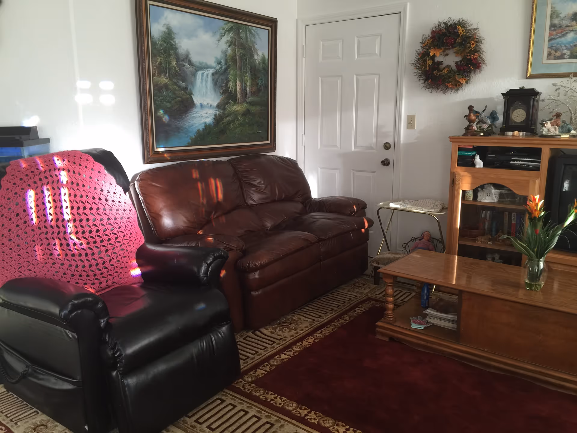 Cozy living room with a brown leather sofa and black recliner, wooden coffee table, entertainment cabinet, and wall artwork.