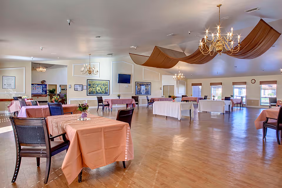 Spacious dining/activity room with multiple tables covered in pink and white tablecloths, chandeliers, and decorative ceiling drapery.