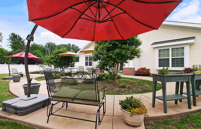 Outdoor patio area with a large red umbrella shading a metal bench with green cushions. There are additional umbrellas and seating areas on a curved concrete pathway surrounded by grass, plants, and a tree. A light-colored building with windows and flower beds is visible in the background under a partly cloudy sky.