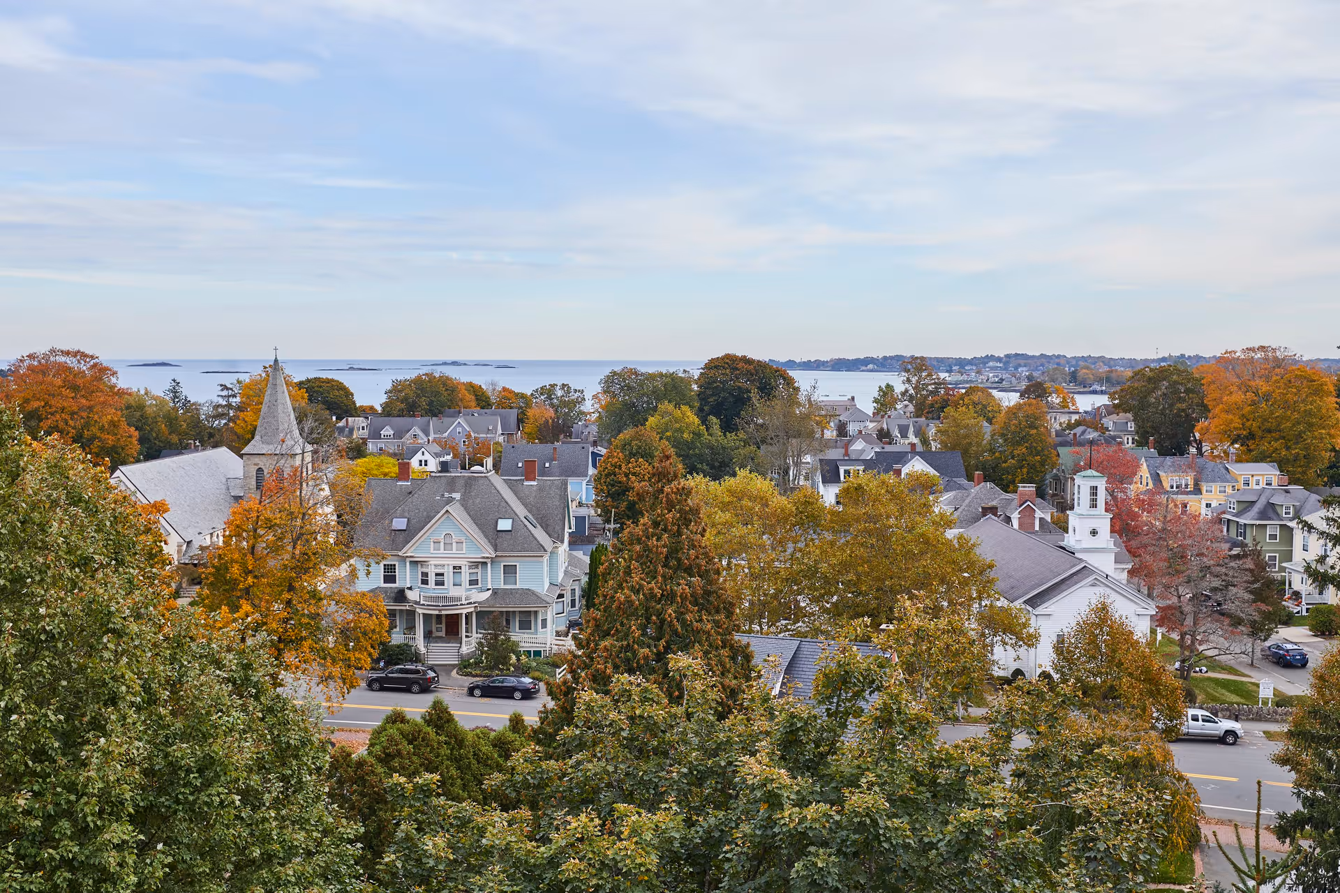 A scenic view of a small town with houses, trees with autumn foliage, a church with a steeple, and a body of water in the background under a partly cloudy sky.