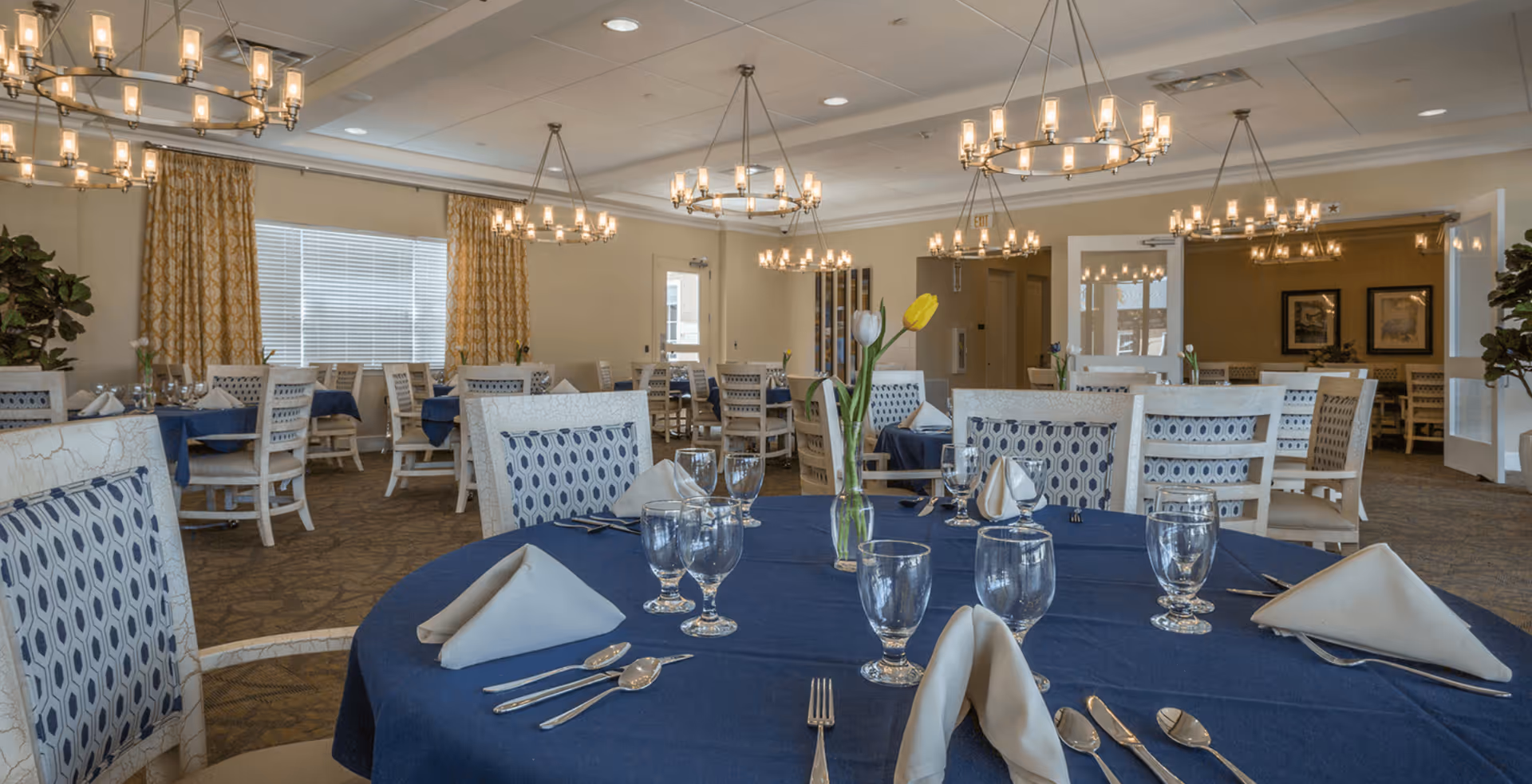 Dining room with round tables set with navy tablecloths, glassware, napkins, and chandeliers overhead.