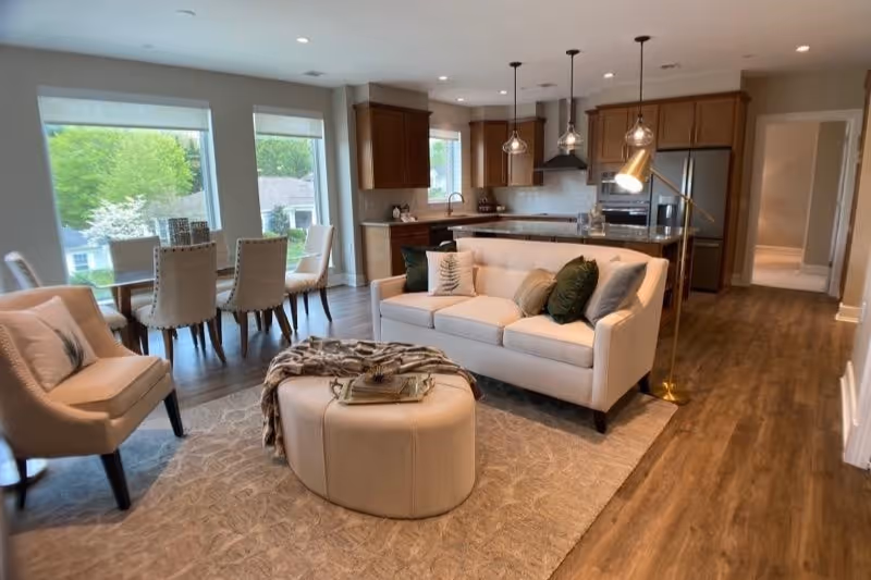 A modern open-concept living room and kitchen area with a beige sofa adorned with green and beige pillows, a round beige ottoman with a throw blanket and books on top, two beige armchairs, a dining table with six upholstered chairs near large windows, wooden flooring, and a kitchen with wooden cabinets, stainless steel appliances, and pendant lighting.