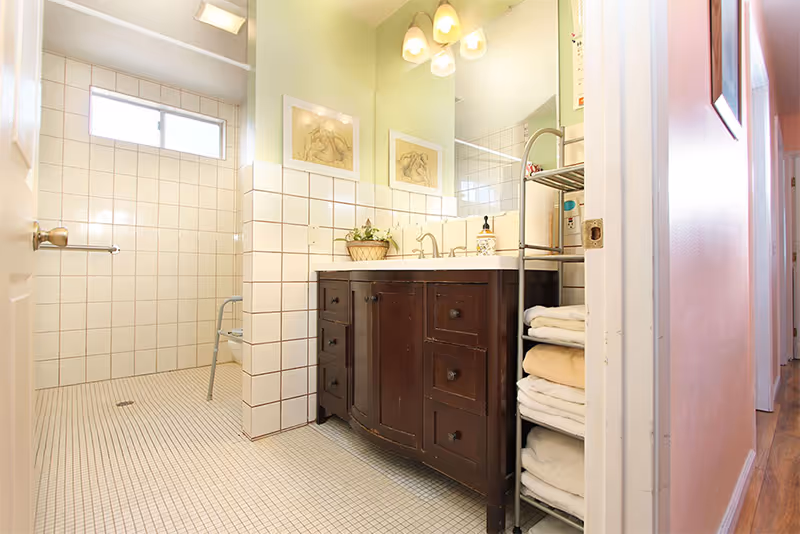 Bright bathroom with a tiled walk-in shower, dark wood vanity with sink and mirror, and a shelf of folded towels.