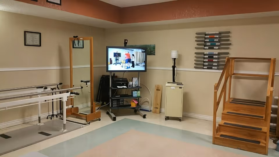 A rehabilitation room with parallel bars for walking exercises, a full-length mirror on a wooden stand, a TV on a rolling cart displaying a person exercising, a wooden set of stairs with handrails, and various therapy equipment mounted on the wall.