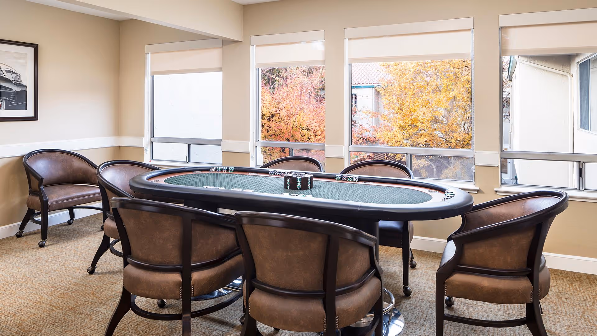 A well-lit room with a poker table surrounded by eight brown leather chairs on casters. The room has beige walls, carpeted floor, and three large windows showing autumn-colored trees outside. A framed black and white photo of a classic car hangs on the left wall.