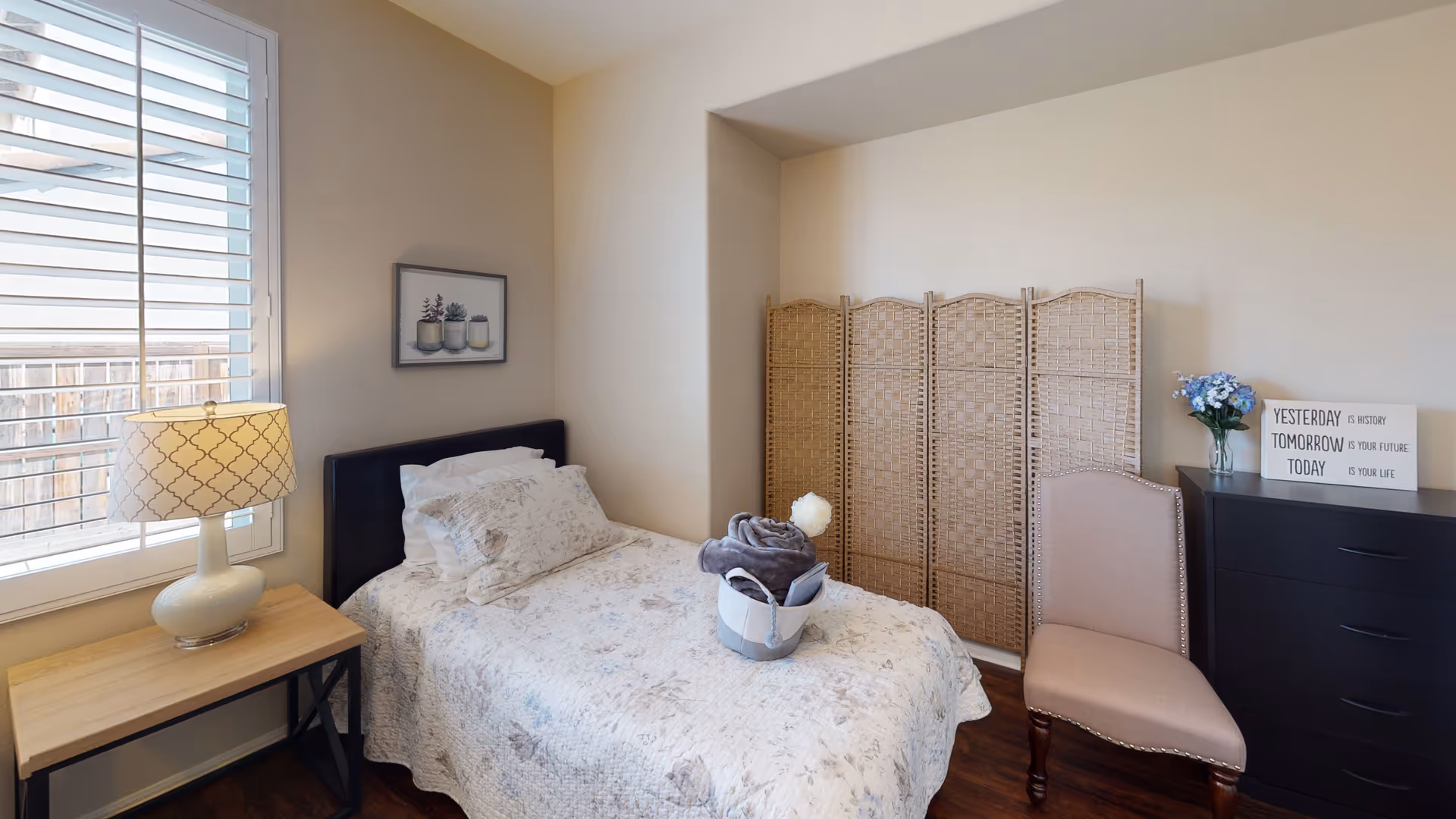 A cozy bedroom with a single bed covered in a floral quilt and pillows. Next to the bed is a wooden side table with a patterned lamp. A window with white shutters allows natural light into the room. On the wall above the bed is a framed picture of potted plants. Across from the bed is a beige upholstered chair and a dark dresser with a vase of blue flowers and a decorative sign that reads 'YESTERDAY IS HISTORY TOMORROW IS YOUR FUTURE TODAY IS YOUR LIFE'. A woven room divider is placed in the corner.