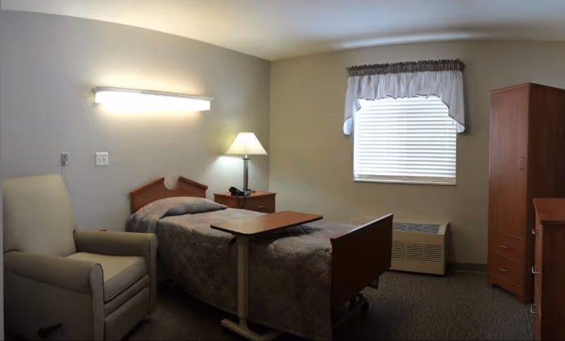 A simple senior care facility bedroom with a single bed covered in a quilted blanket, a beige armchair, a wooden nightstand with a lamp and telephone, a window with blinds and a valance, a wooden wardrobe, and a small overbed table.