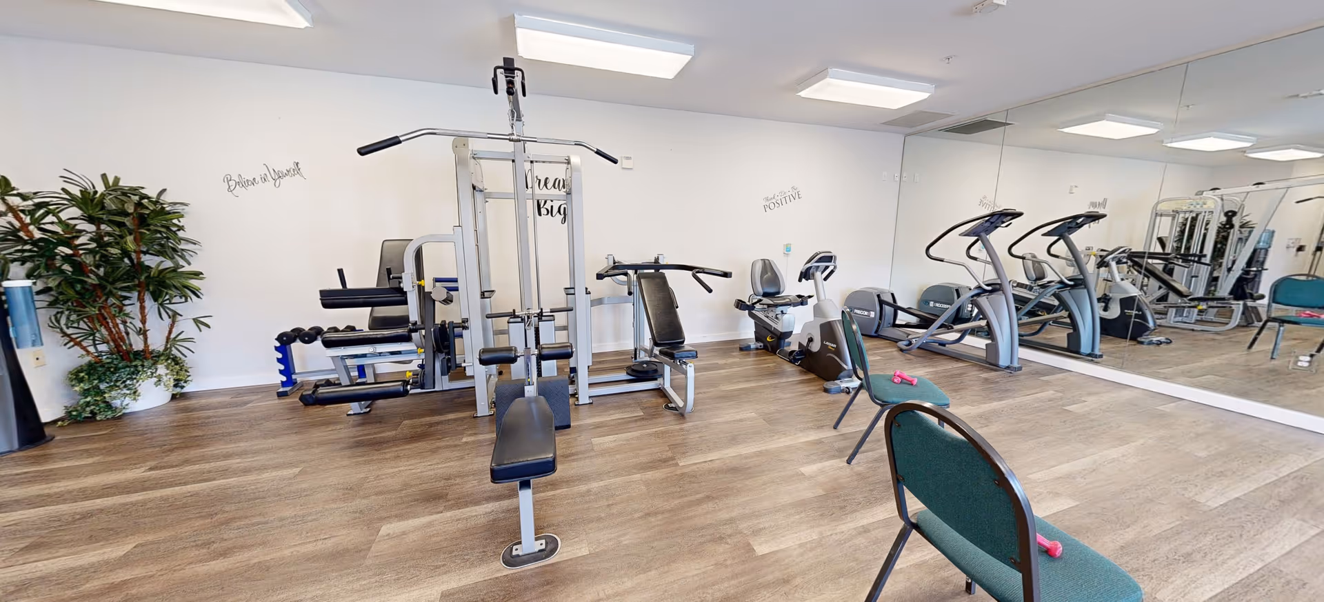 A fitness room with exercise equipment including a multi-station weight machine, two recumbent bikes, and two elliptical machines. The room has wood flooring, white walls with motivational quotes, a large mirror on one wall, and two green chairs with pink dumbbells placed on them. There is also a potted plant in the corner.