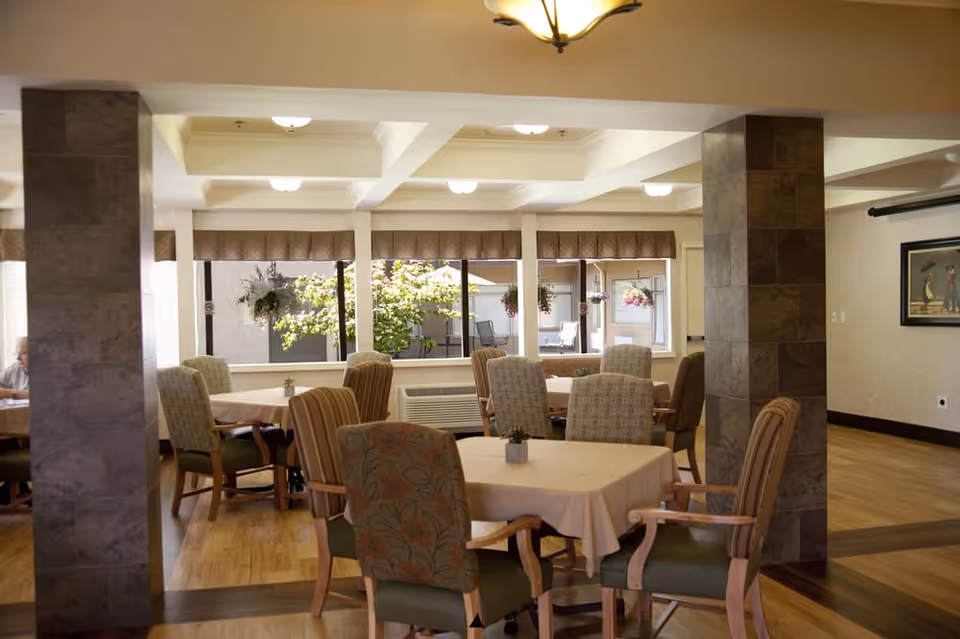 A bright dining/common room with several tables and upholstered chairs, tiled columns, and large windows overlooking hanging plants.