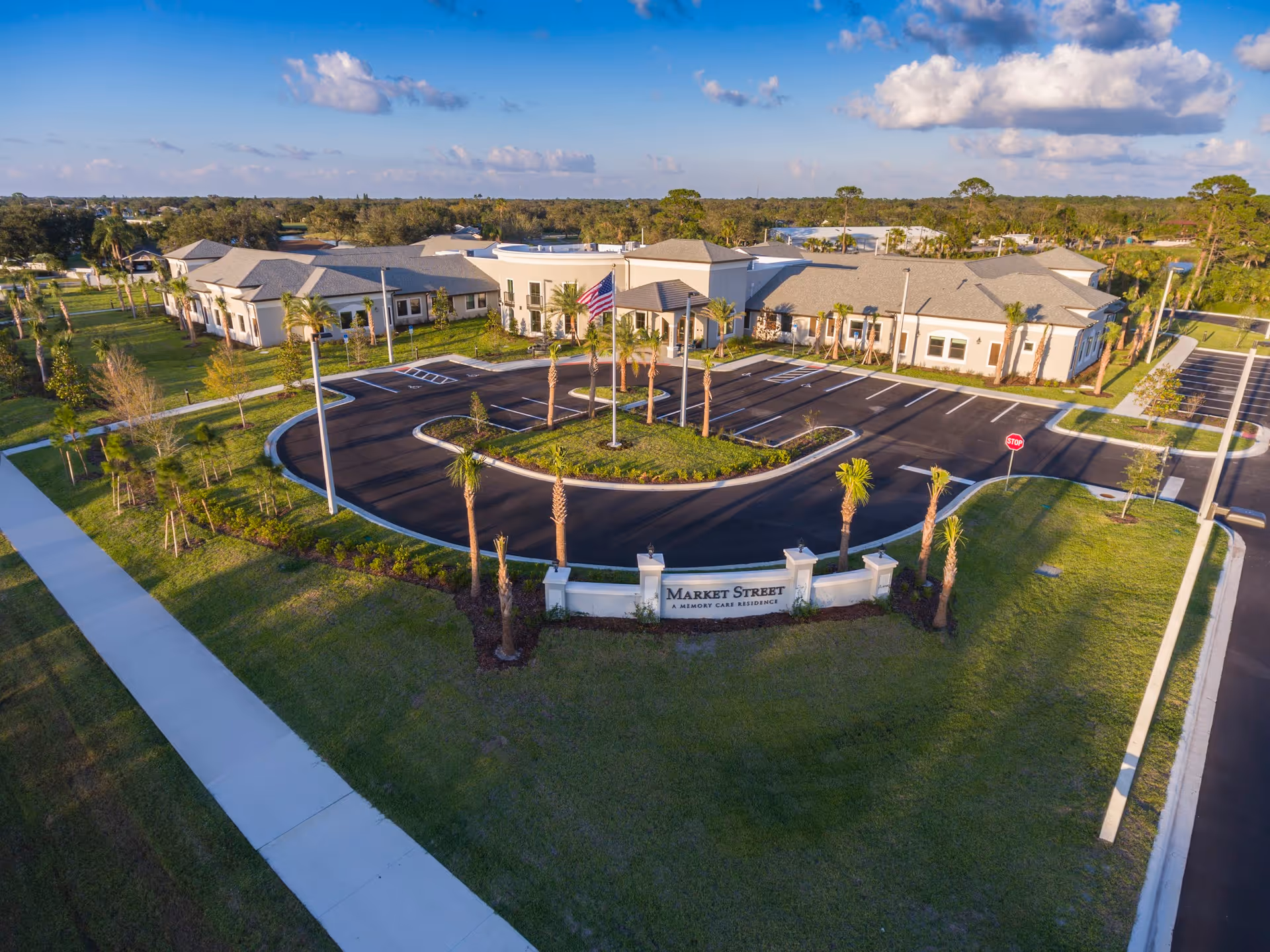 Aerial view of Market Street Viera, a memory care residence, showing a large single-story building with a circular driveway and parking lot surrounded by green lawns and palm trees under a partly cloudy sky.