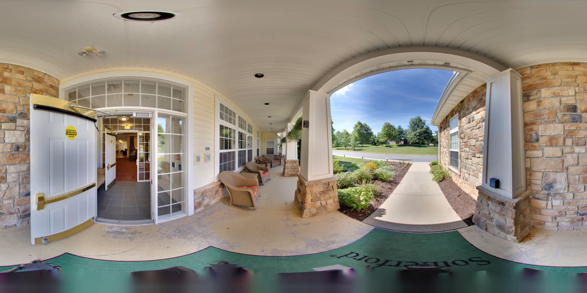 Covered entrance porch of Somerford House Frederick with stone pillars and beige siding. There are several cushioned chairs along the wall, a green welcome mat on the floor, and a pathway leading to a landscaped garden and a road. The entrance door is open, showing a glimpse of the interior with tiled flooring.