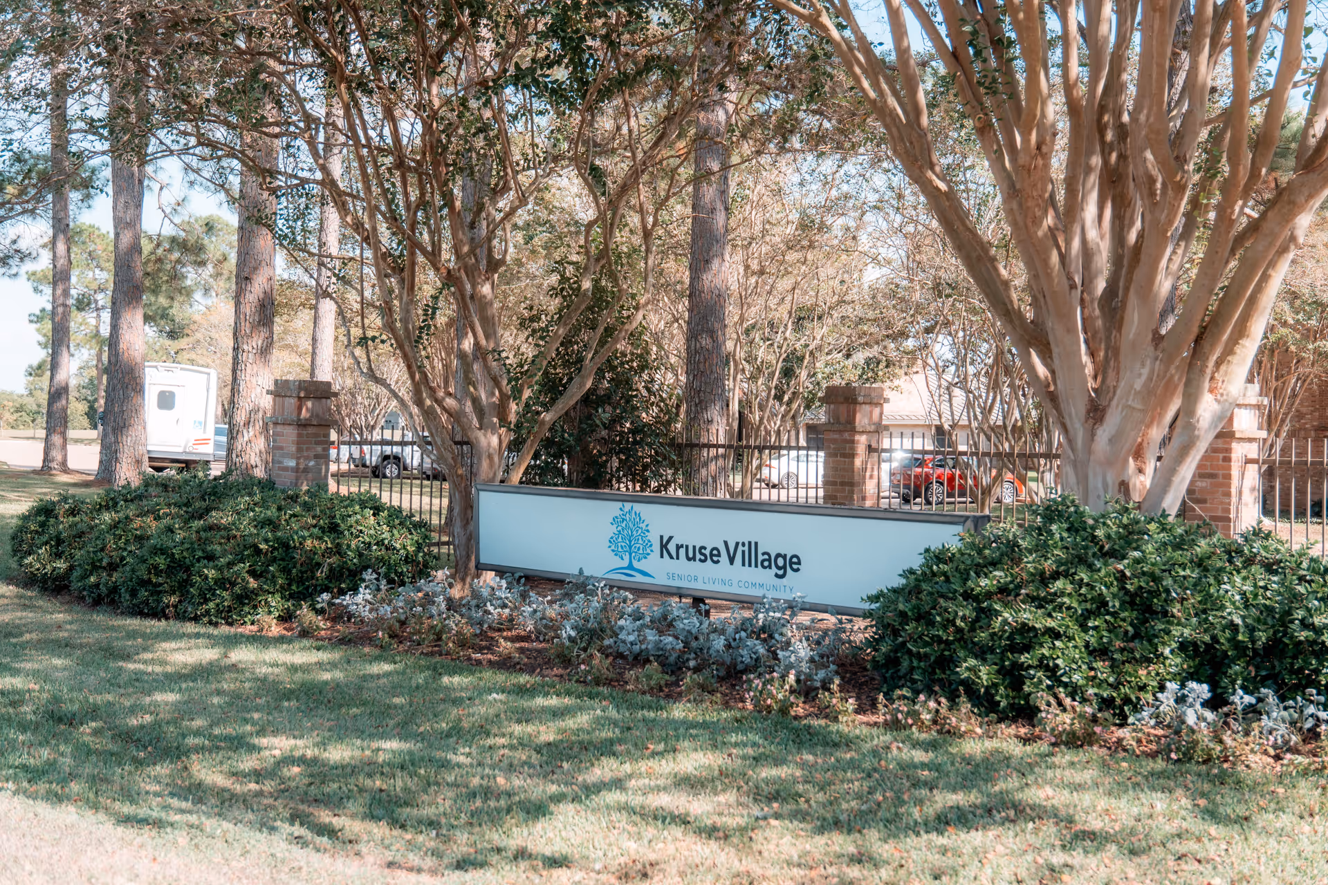 Outdoor view of the entrance sign for Kruse Village Senior Living Community surrounded by trees, bushes, and grass with a parking area and vehicles visible in the background.