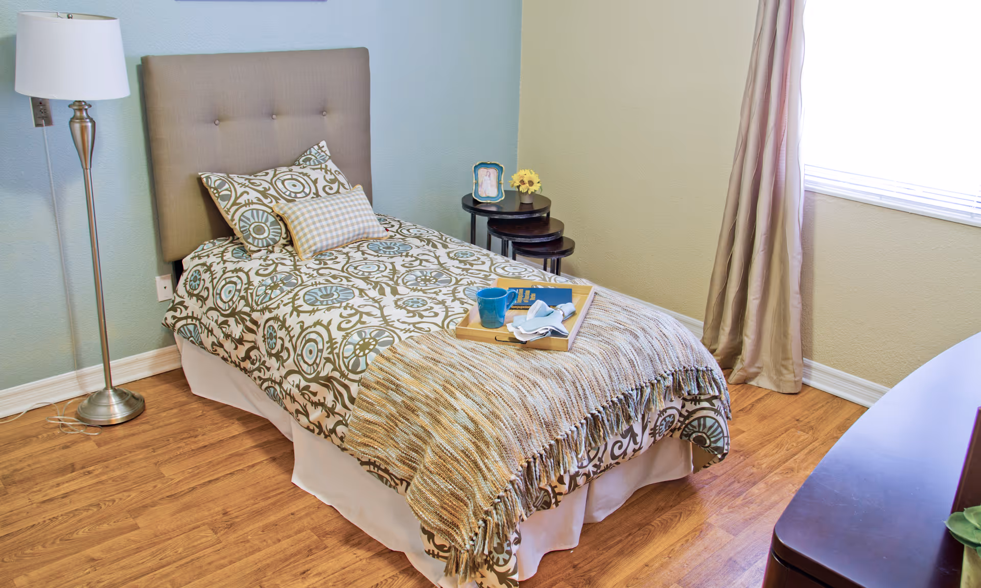 Single bed with patterned bedding, a throw and a breakfast tray in a bright, simply furnished bedroom with a floor lamp and window.