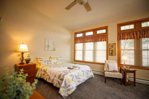 A cozy bedroom with a bed covered in a floral patterned comforter, a wooden nightstand with a lamp and small plant, a cushioned chair next to a small wooden table with a lamp, and large windows with blinds and valances letting in natural light.