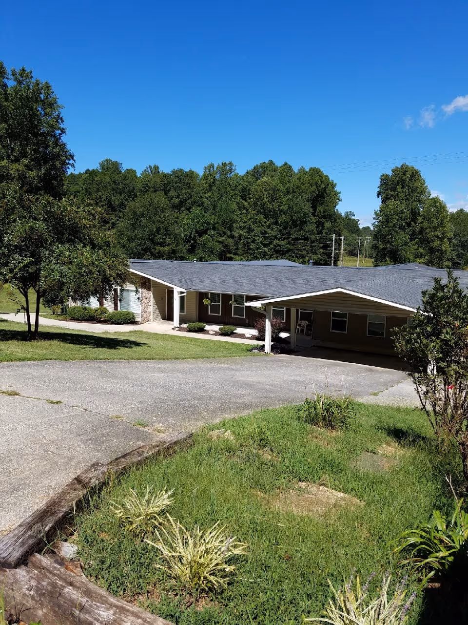 Single-story building with a sloped roof surrounded by green grass, trees, and a paved driveway under a clear blue sky.