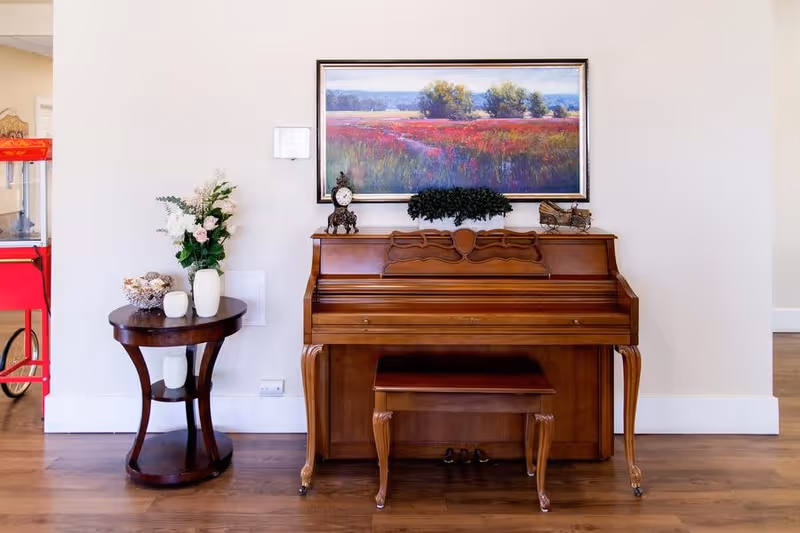 A wooden upright piano with a matching bench is placed against a white wall. Above the piano hangs a large framed painting of a colorful field with trees in the background. On top of the piano are decorative items including a clock, a small plant, and a miniature cart. To the left of the piano is a round wooden side table with a vase of flowers and other decorative objects. The floor is wooden, and part of a red popcorn machine is visible on the far left.