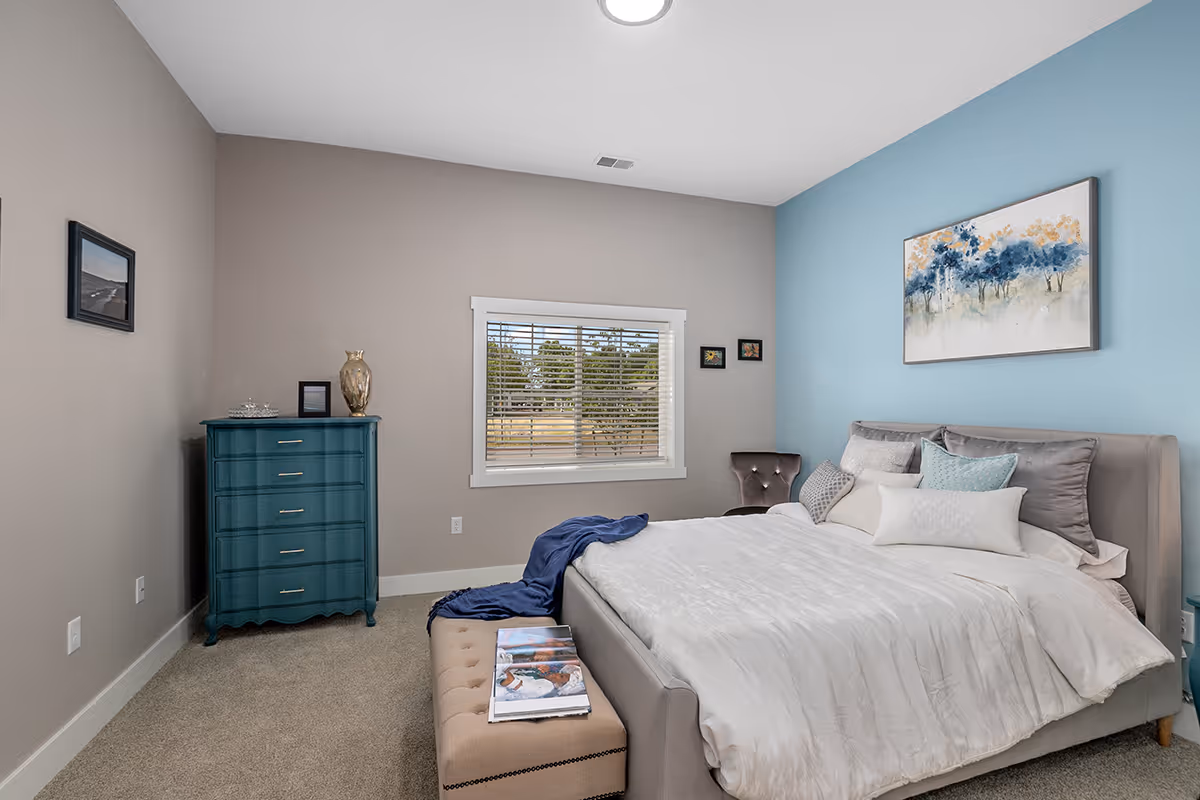 A neatly made bed with multiple pillows and a white comforter in a bedroom with light blue and beige walls. There is a beige cushioned bench at the foot of the bed with magazines on it. A teal chest of drawers with decorative items on top is against one wall, and a window with white blinds shows trees outside. Several framed pictures hang on the walls.