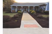 Front exterior view of a single-story building with a covered porch, multiple columns, and a concrete walkway leading to the entrance, surrounded by landscaped bushes and mulch beds.