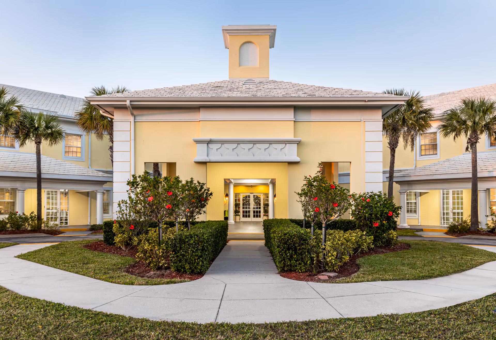 Front entrance of a yellow two-story assisted living building with symmetrical landscaping, palm trees, and a paved walkway.
