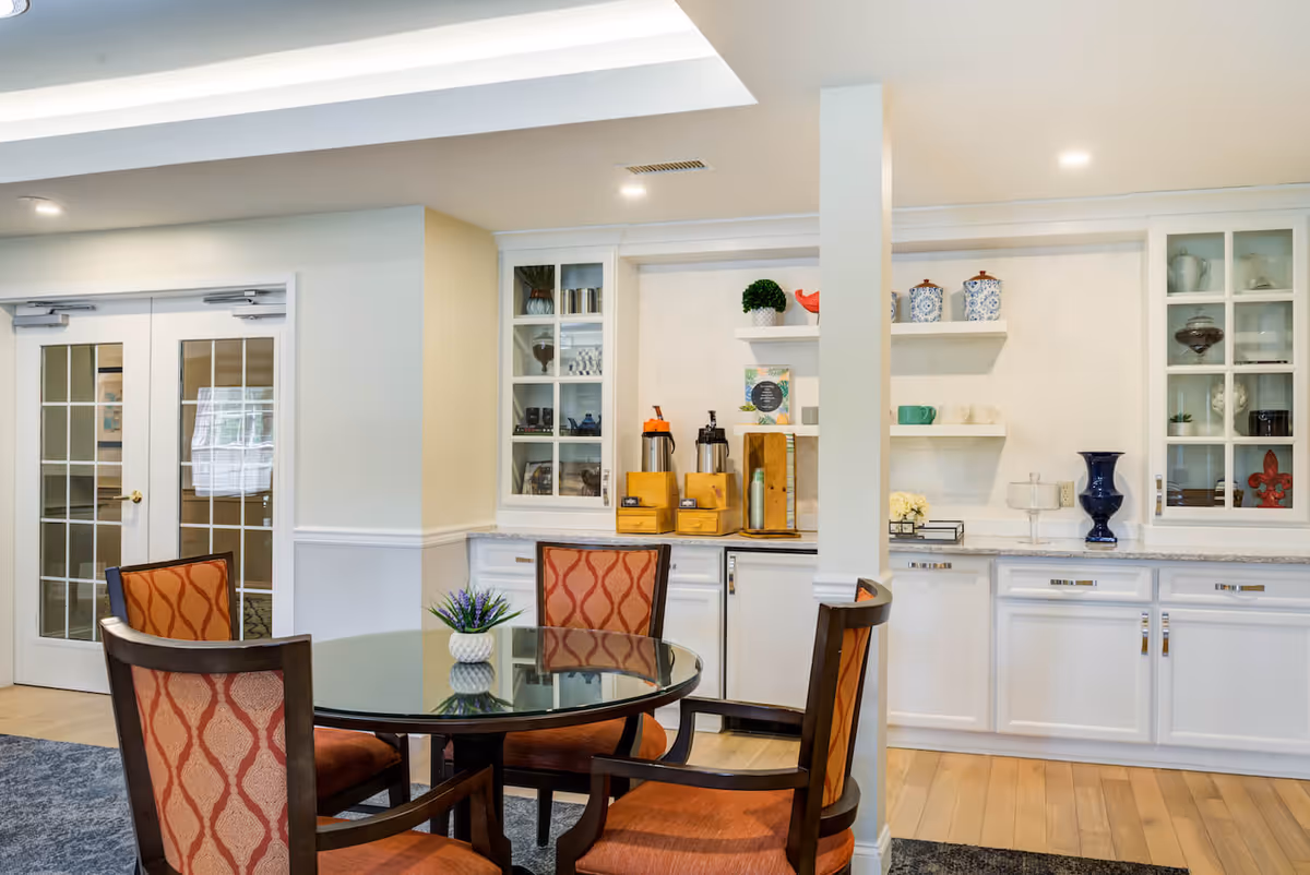 A cozy interior space featuring a round glass table surrounded by four upholstered chairs with orange patterned fabric. Behind the table is a built-in white cabinet with glass doors displaying decorative items and shelves holding coffee dispensers, cups, and ornamental pieces. The room has light-colored walls, wooden flooring, and double glass doors in the background.