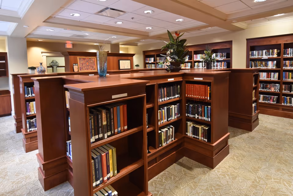 Interior view of a library with wooden bookshelves filled with books and potted plants on top.