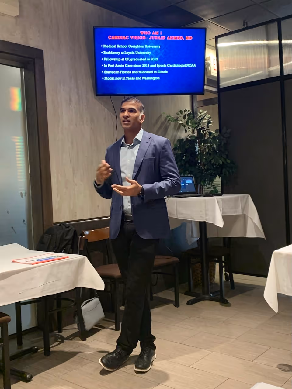 A man in a blue blazer and black pants is giving a presentation in a room with tables covered in white tablecloths. Behind him, a screen displays a slide titled 'WHO AM I CARDIAC VISION - JUMAID AHMED, MD' with bullet points about his medical education and career. There is a laptop on a table in the background and a plant next to it.