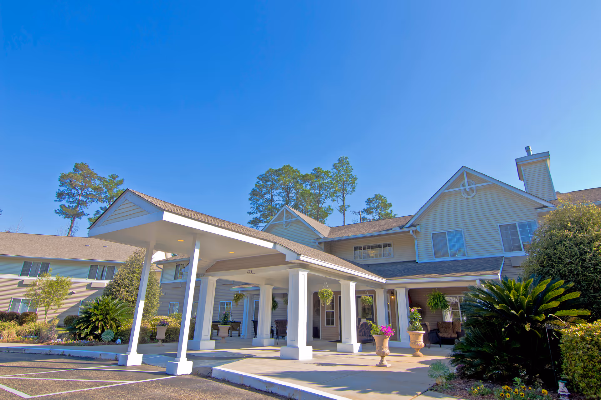 Exterior view of Brookdale Hattiesburg senior living facility showing the main entrance with a covered drop-off area supported by white columns, surrounded by well-maintained landscaping and greenery under a clear blue sky.