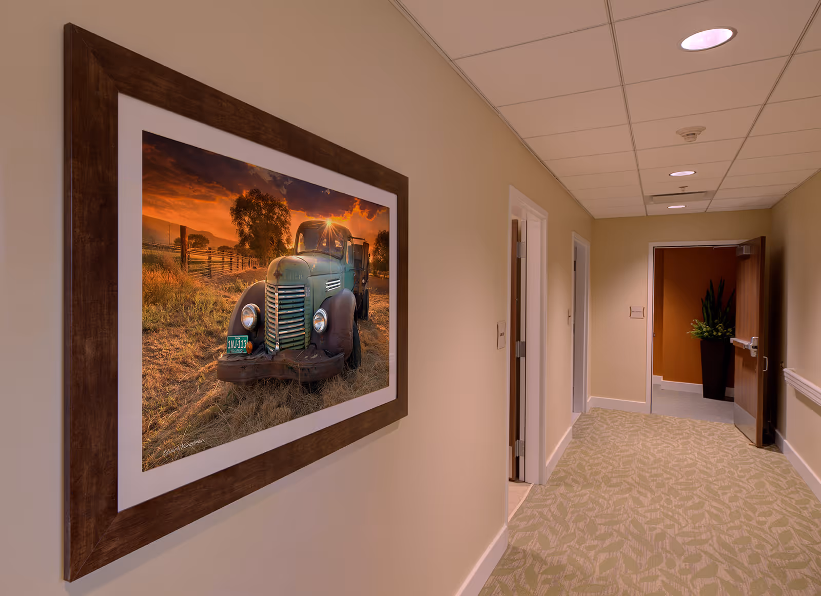 A hallway in The Lodge at Grand Junction with beige walls and patterned carpet. On the left wall, there is a large framed photograph of an old green truck in a field at sunset. The hallway leads to an open door revealing a room with a large plant in a tall black planter.