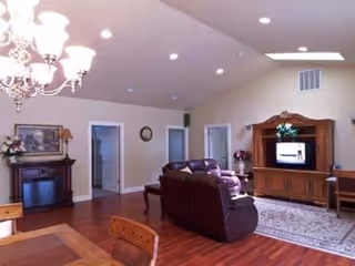 A spacious living room with high ceilings featuring a chandelier, a wooden dining table with chairs in the foreground, a dark leather sofa set facing a wooden entertainment center with a TV, a fireplace with a framed picture above it, and doorways leading to other rooms.