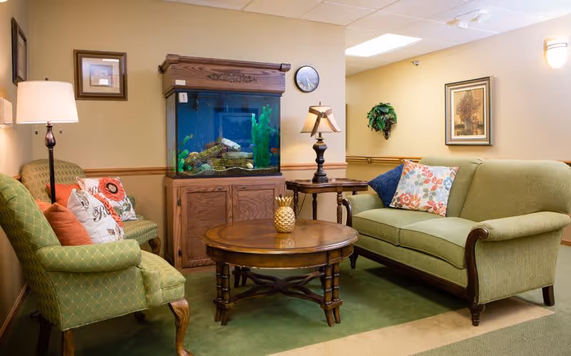 A cozy living room area with a green upholstered sofa and two green armchairs with patterned cushions. There is a wooden coffee table with a decorative pineapple on it, a wooden side table with a lamp, and a large fish tank on a wooden cabinet. The walls are beige with framed artwork and a wall clock.