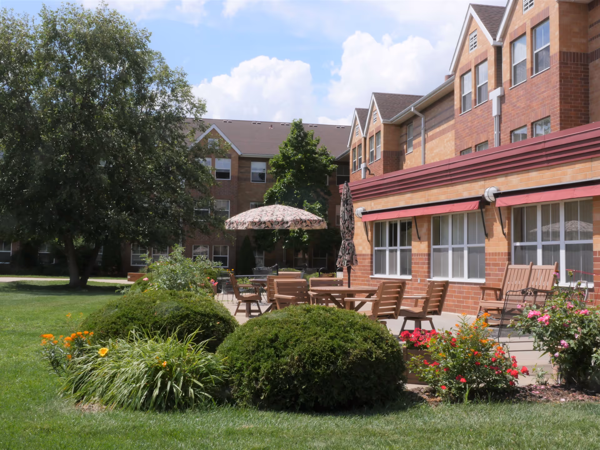 Outdoor patio area of a senior living facility with wooden tables and chairs, umbrellas, and well-maintained bushes and flowers in the foreground. The building has a brick exterior with multiple windows and a red awning. A large tree and a partly cloudy sky are visible in the background.