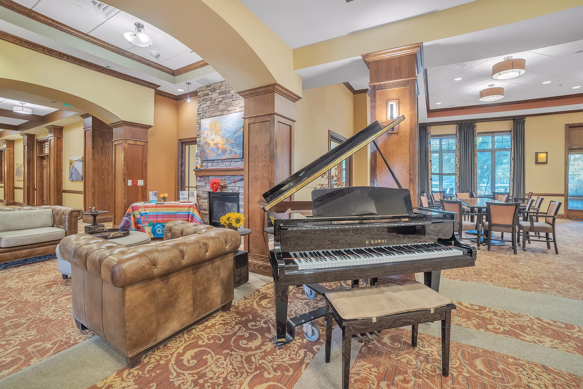 A spacious and warmly decorated common area featuring a black grand piano with a bench, leather sofas, a stone fireplace with artwork above it, and a dining area with tables and chairs near large windows with curtains.