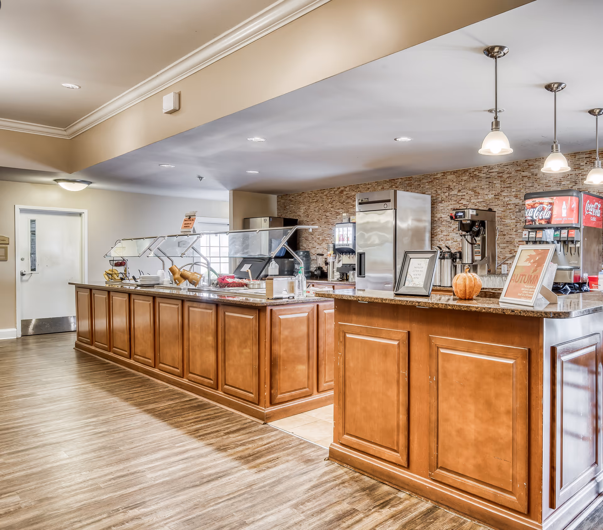 Interior view of a dining service area with a long wooden counter featuring a glass sneeze guard, plates, and serving utensils. Behind the counter are beverage dispensers including a Coca-Cola machine, coffee makers, and a refrigerator. The floor is wooden, and the ceiling has recessed lighting and hanging pendant lights. There are framed signs and a small pumpkin on the counter, suggesting a seasonal decoration.