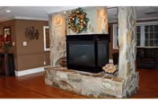 A cozy interior common area featuring a central stone fireplace with a decorative wreath above it. The room has wooden flooring and neutral-colored walls, with seating areas visible in the background.