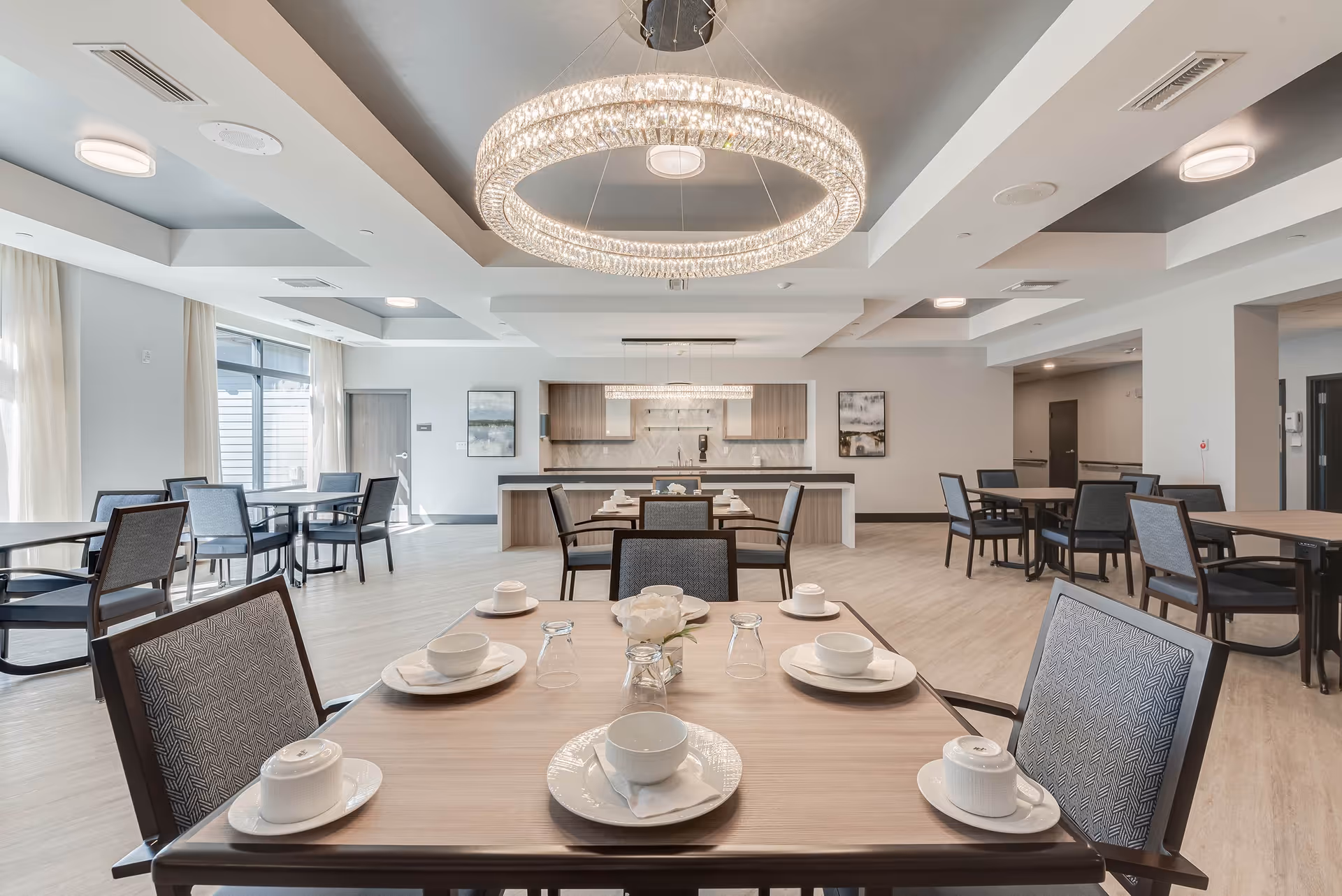 Modern communal dining room with set tables, a large circular chandelier, and a serving kitchen in the background.