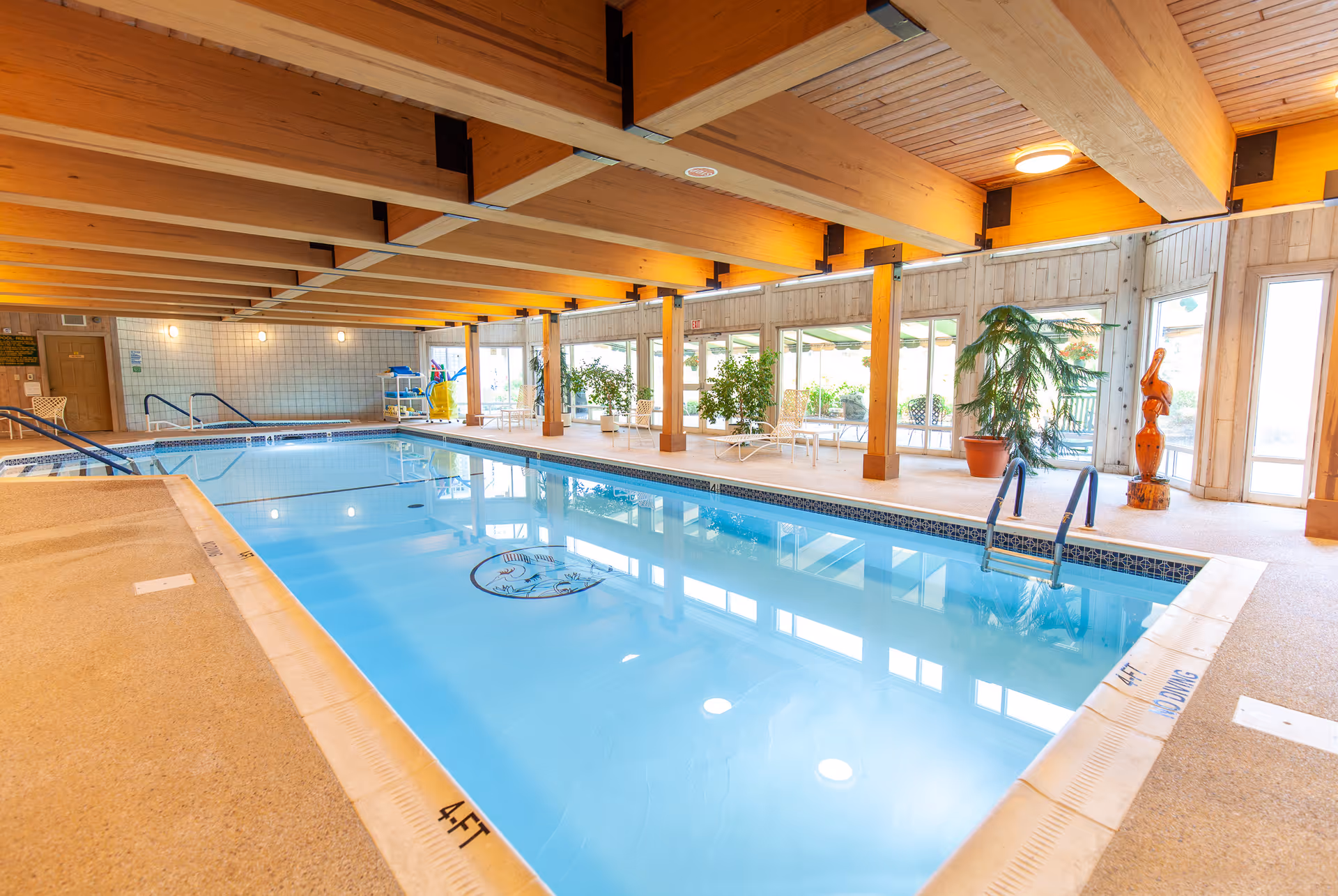 Indoor swimming pool room with wooden ceiling beams, large windows, plants and poolside seating.
