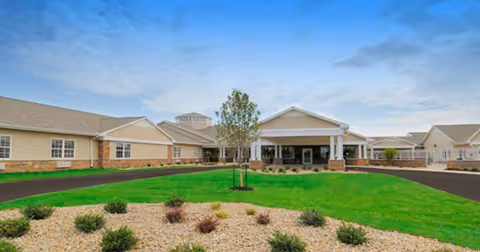 Front exterior view of a single-story senior living facility building with a covered entrance, surrounded by a well-maintained lawn, small shrubs, and a clear blue sky.