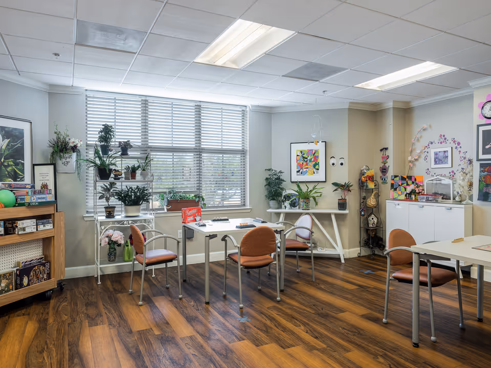 A bright and cozy activity room with wooden flooring, several tables and chairs arranged for games or crafts, shelves with board games, and various potted plants and colorful artwork decorating the walls.