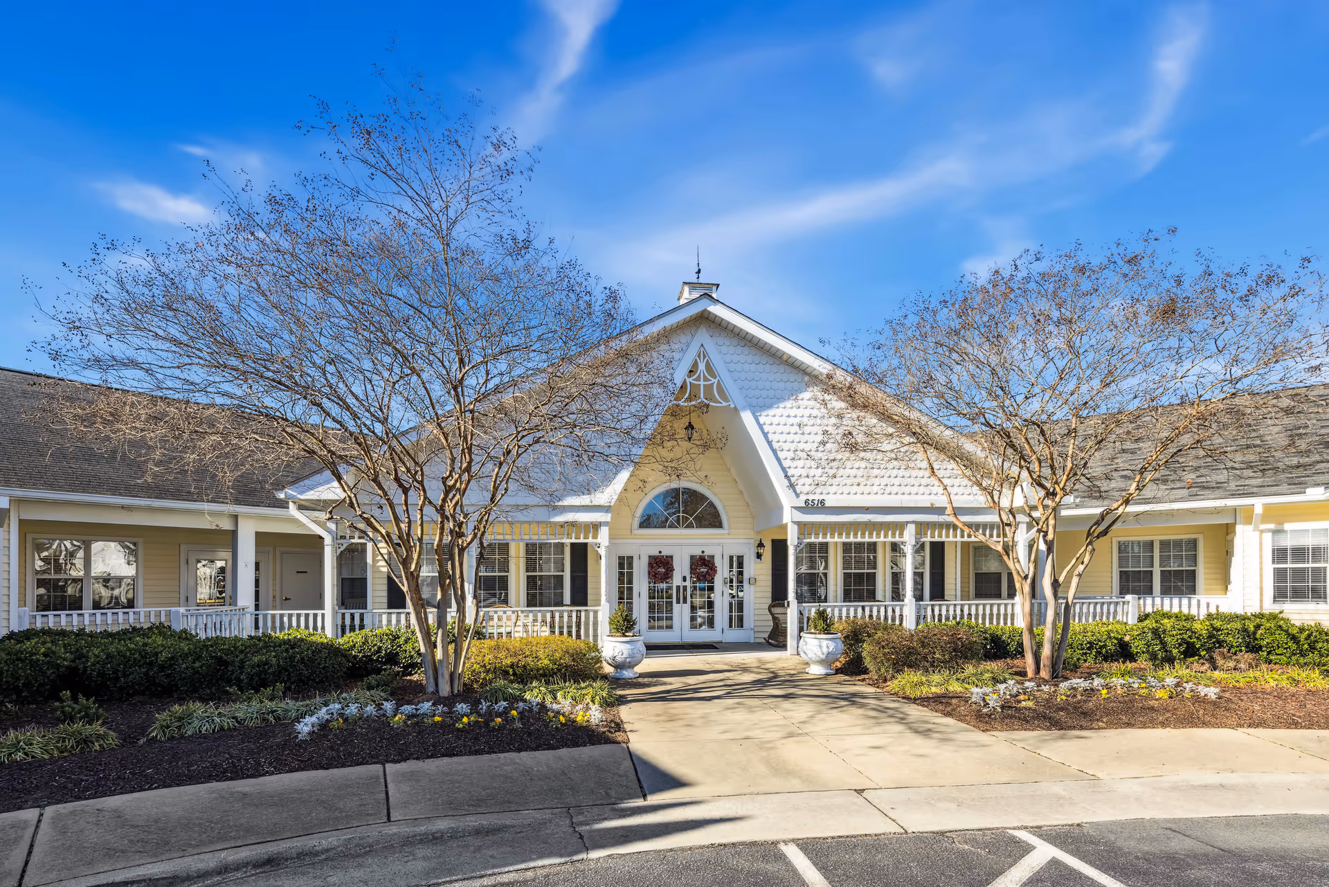 Front exterior view of a single-story senior living facility building with a peaked roof, white siding, and large windows. There are two leafless trees and landscaped bushes and flowers in front of the building, with a clear blue sky above.
