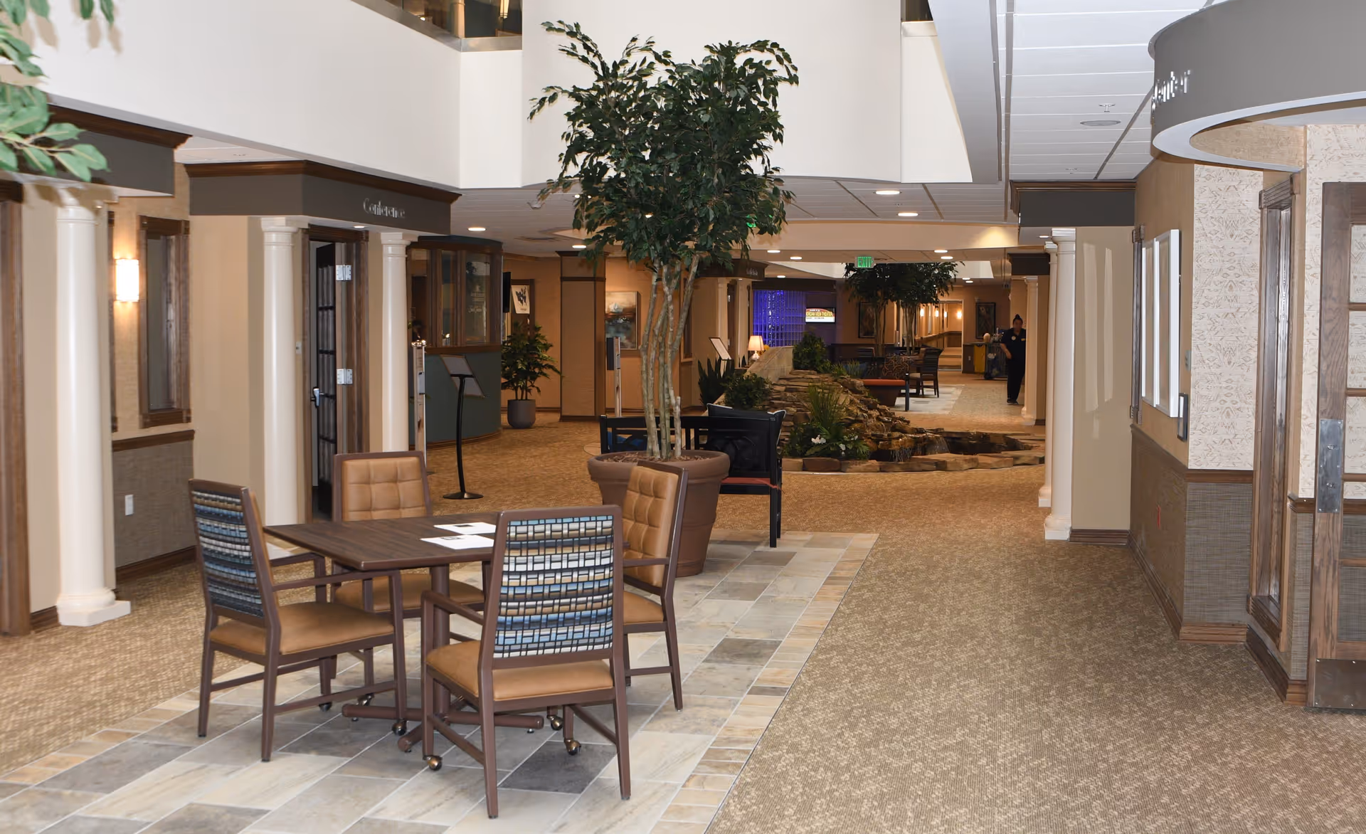 Interior view of a retirement community hallway with a seating area featuring a square table and four chairs on a tiled floor section. The hallway has beige carpet, potted plants including a large tree, decorative columns, and warm lighting. A water feature with rocks and plants is visible further down the corridor, along with additional seating and a person walking in the background.