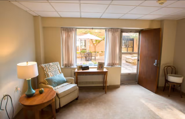 A cozy sitting room with an upholstered armchair, side tables and lamp beside a window and an open door leading to an outdoor courtyard.
