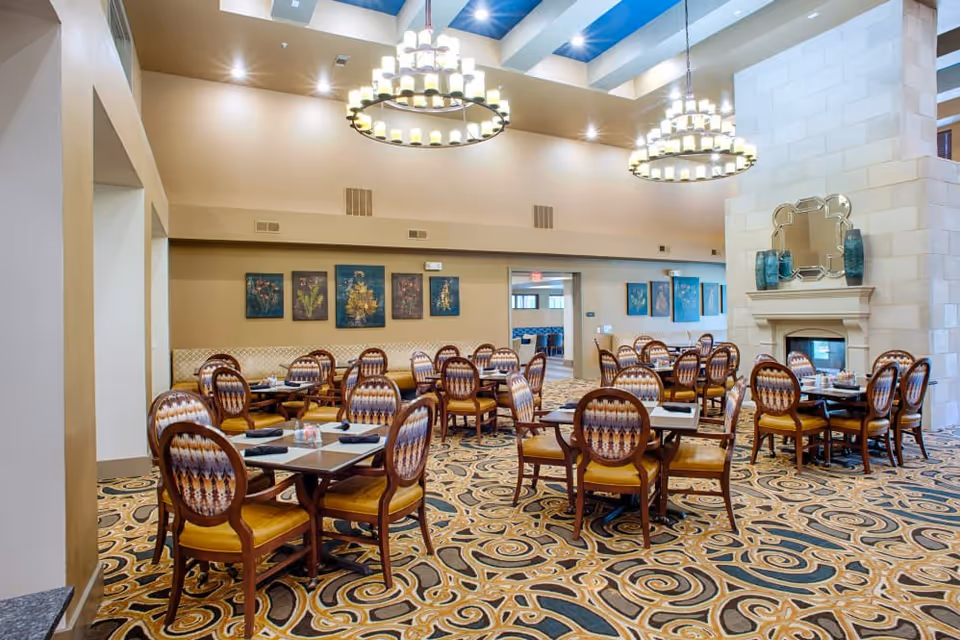 A spacious dining room with multiple wooden tables and chairs arranged neatly. The chairs have patterned upholstery and yellow cushions. The room features a large decorative carpet with intricate designs, a stone fireplace with a mirror and vases on the mantel, and two large circular chandeliers hanging from the ceiling. The walls are adorned with framed artwork and the ceiling has skylights allowing natural light to enter.