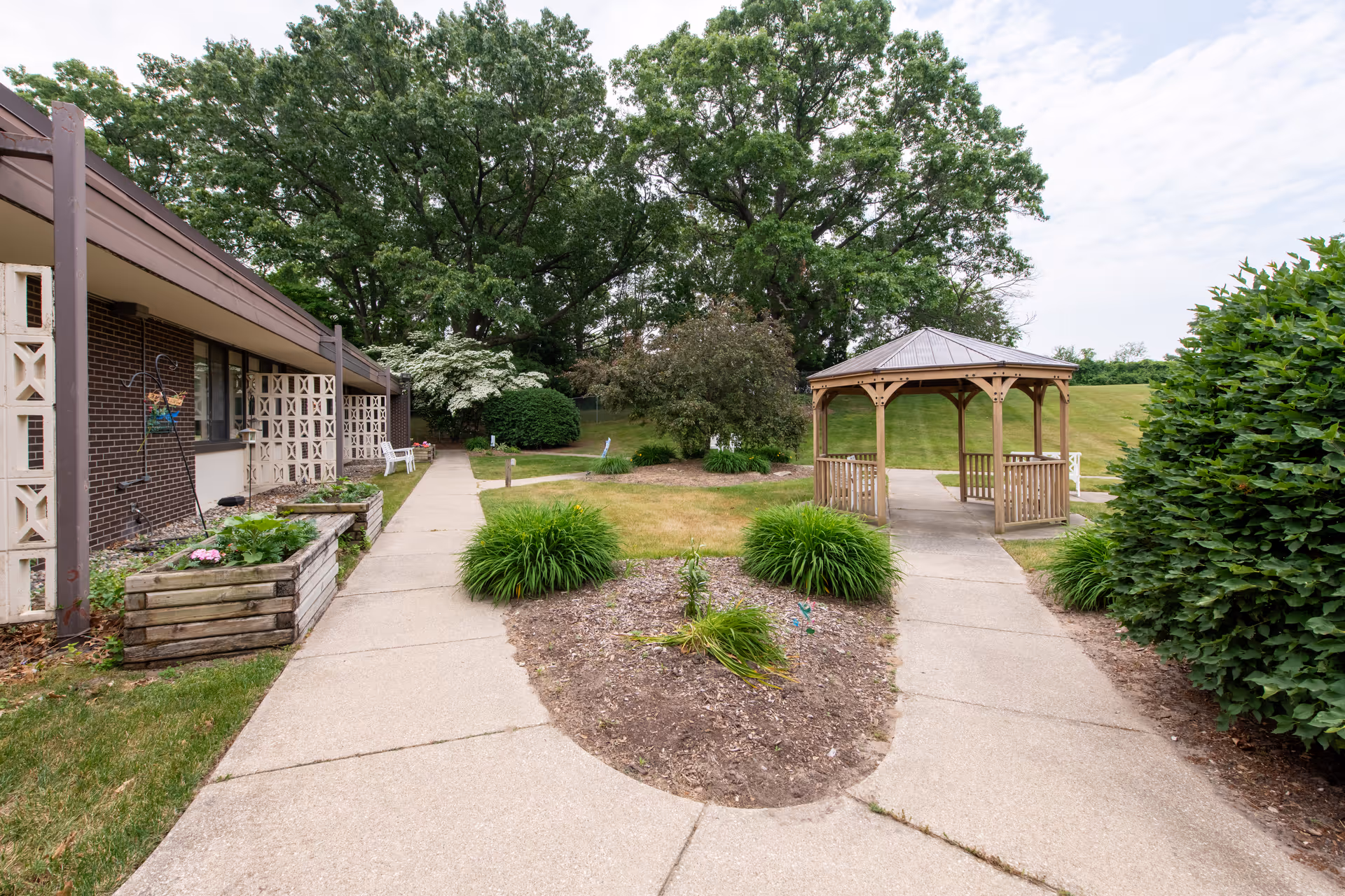 Outdoor garden area at SKLD Leonard featuring a paved walkway splitting into two paths around a small garden bed with green plants. There is a wooden gazebo on the right side and a brick building with decorative concrete blocks on the left. Large trees and bushes surround the area under a partly cloudy sky.